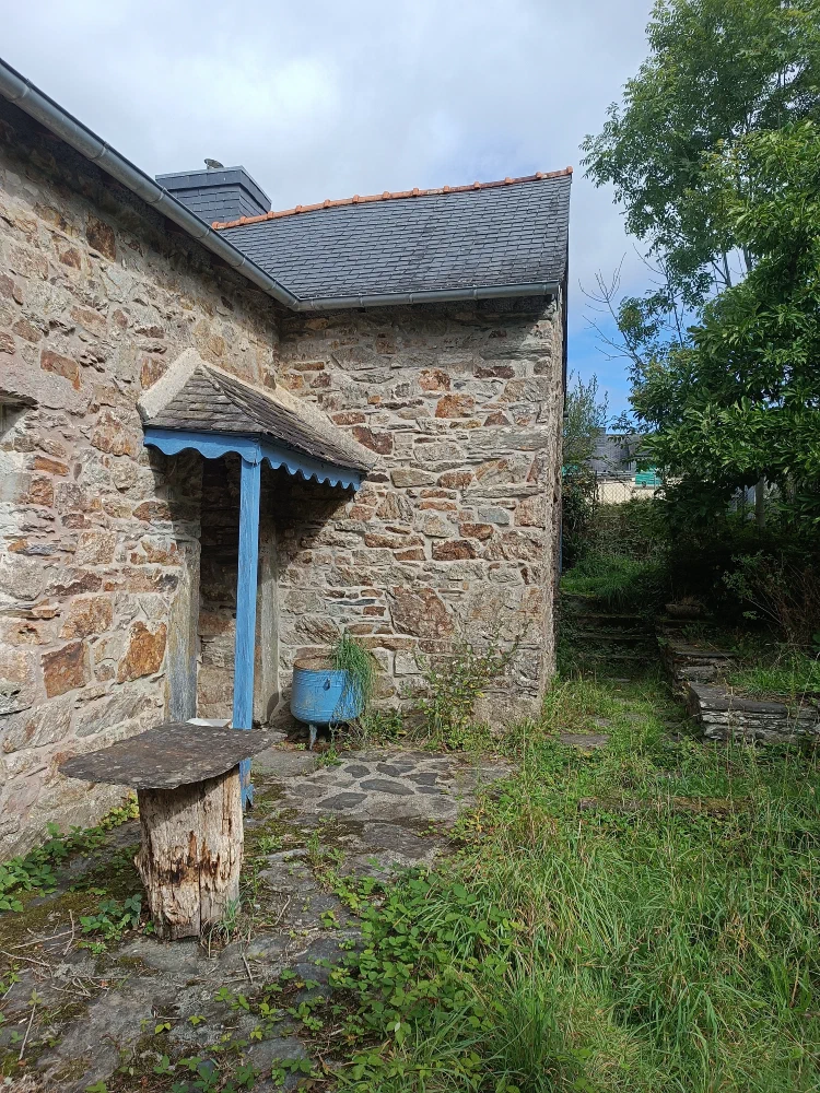 Le pignon en pierre de la maison, avec son auvent bleu au-dessus de la porte, un banc de pierre posé sur un rondin et le jardin qui monte vers l'arrière