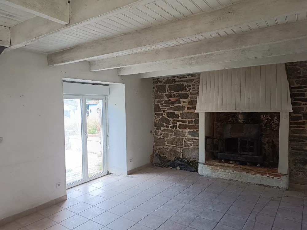 Le séjour vide aux poutres blanches, avec sa grande cheminée et sa porte-fenêtre ouvrant sur l'extérieur