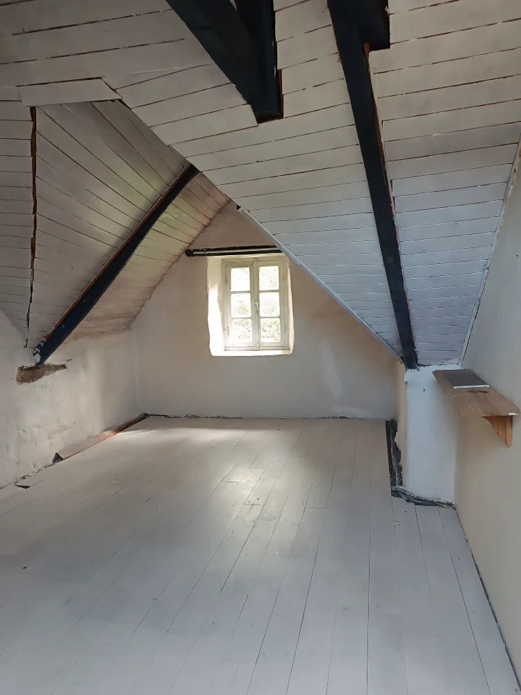 Une chambre à l'étage au plancher peint en blanc, avec ses poutres sombres et sa petite fenêtre à croisillons