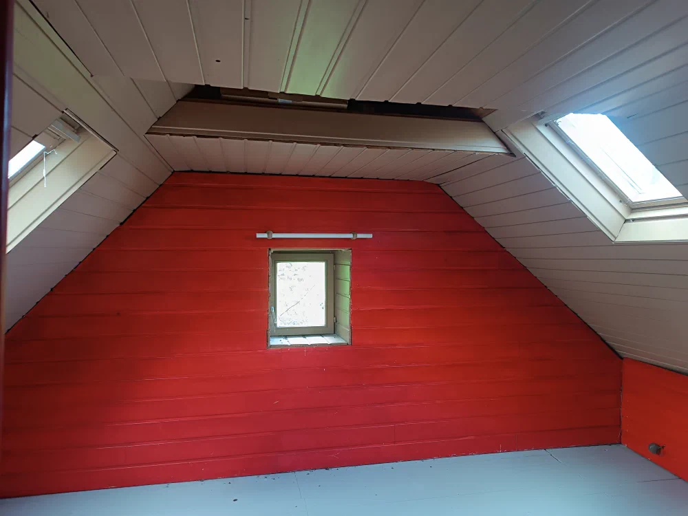 Une pièce mansardée aux murs rouges, éclairée par deux fenêtres de toit et une petite lucarne