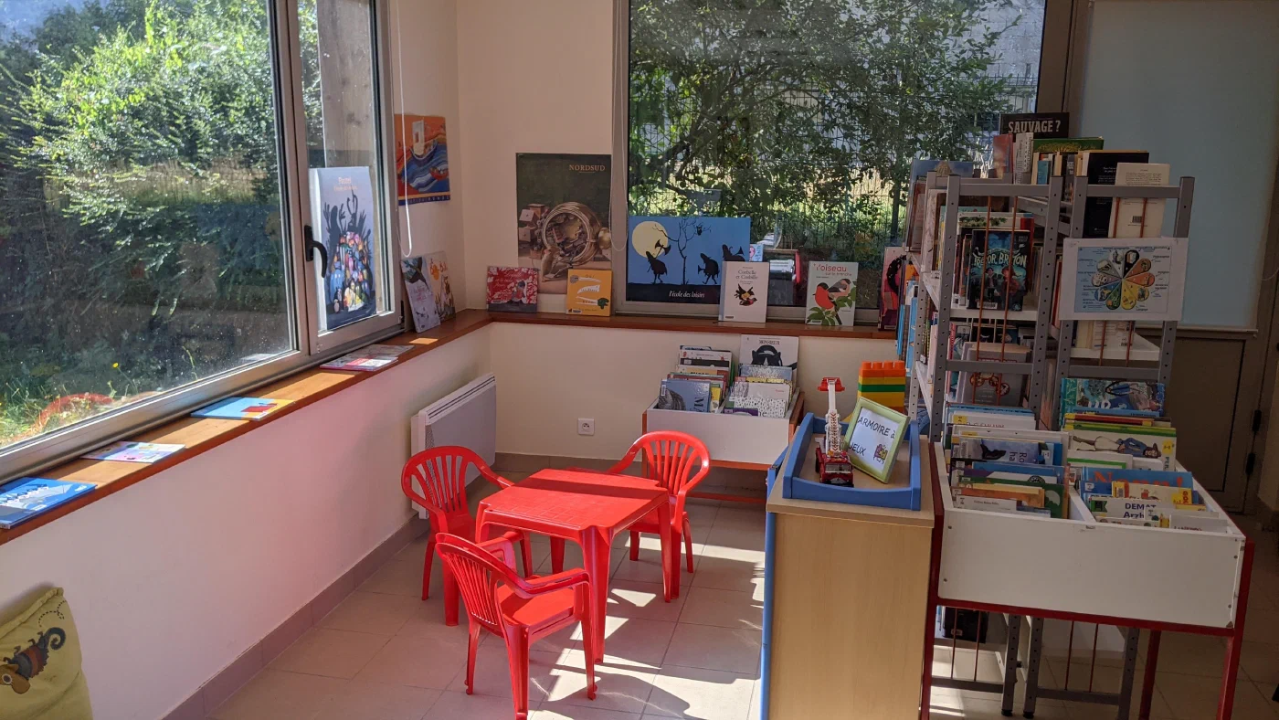 Le coin jeunesse de la bibliothèque, avec sa table et ses chaises rouges et ses albums exposés devant les fenêtres